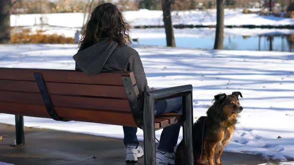 Man with dark long hair sitting on a bench with his dog at a park in Winter
