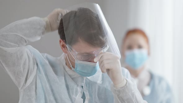 Concentrated Male Doctor Putting on Face Shield with Blurred Female Nurse in Coronavirus Face Mask