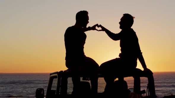 Happy caucasian gay male couple sitting on car making heart shape with hands at sunset on the beach