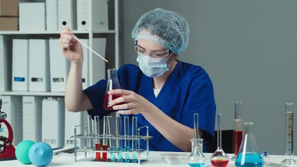 Young Doctor Wearing Medical Protective Mask with Gloves Mixing and Pouring Red Solution Into Flask alt