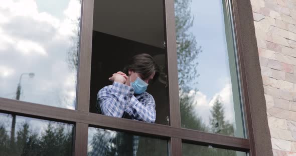 Young Man in Medical Mask Looks Through Open Window and Bored During Isolation