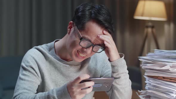 Close Up Of Asian Man Being Upset Losing Game On Smartphone After Working With Documents At Home