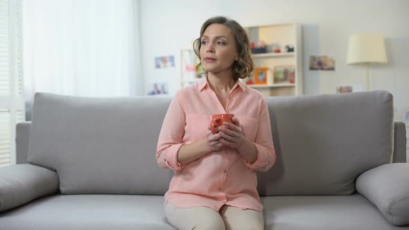 Sad Young Female With Cup of Tea Looking Camera Sitting on Sofa, Remembrance