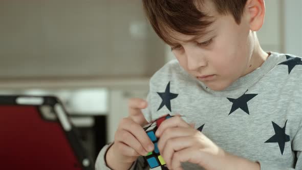 Concentrated boy in grey sweatshirt holding cube and playing with it while watching tablet