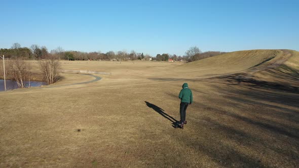 An aerial tracking of a man on an electric skateboard in an empty park on a sunny day. The drone fol