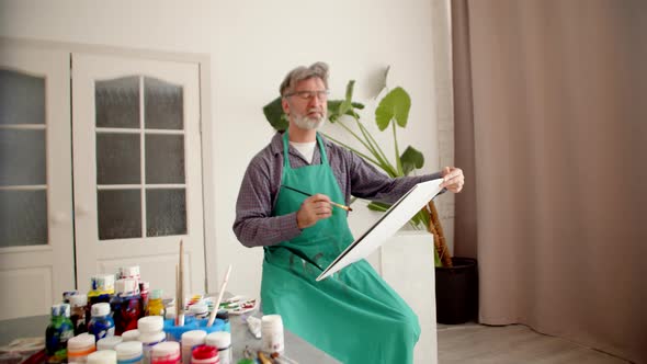 Man with Gray Hair Sitting on Chair Drawing on Canvas with Brush Dipping It in Paint Side View