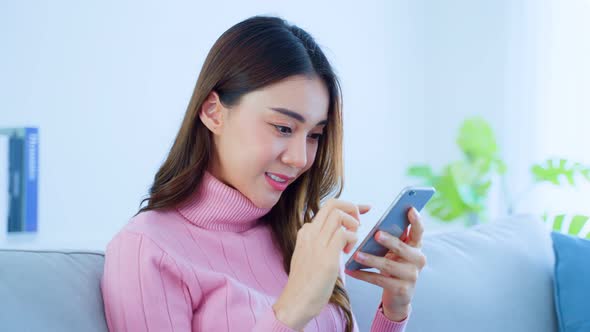 Asian beautiful woman sit on sofa click and swiping on phone enjoy social media in living room.