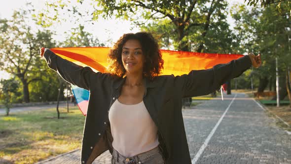 Afroamerican Model is Holding Colorful LGBT Pride Flag Above Her Head Waving It and Smiling While