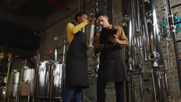 Diverse male and female colleague at gin distillery inspecting equipment, talking and making notes