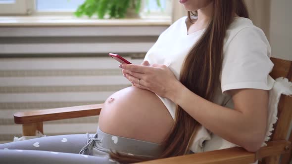 Pregnant Woman Using Phone and Touching Belly While Sitting in Armchair in Home Room Spbd