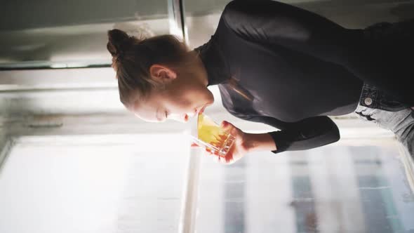 Young woman sitting on a windowsill and enjoying a drink while looking outside on a sunny afternoon