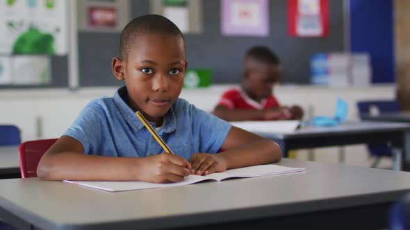 Portrait of happy african american schoolboy sitting at classroom, making notes, looking at camera