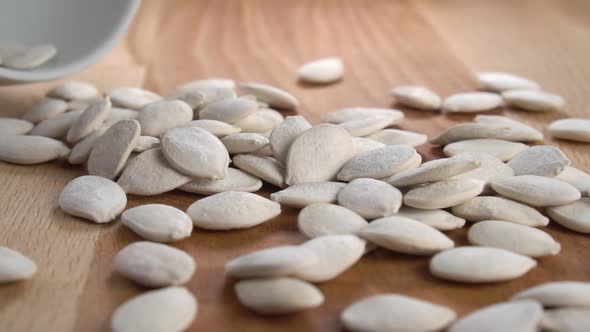 Dried salted pumpkin seeds with shells fall from a white bowl onto a rustic wooden surface