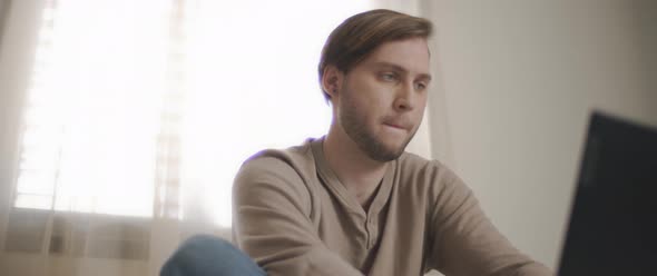 A close up of a young man sitting in bed and working on a computer