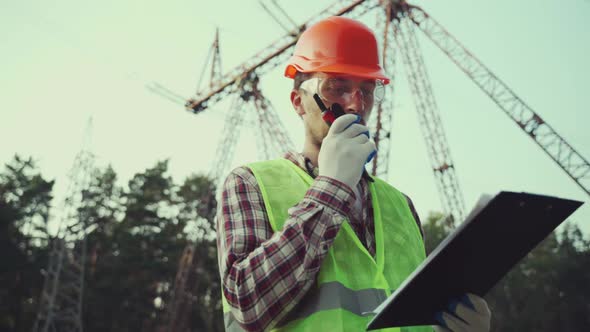 Electrical Equipment Worker Near High Voltage Tower Using Walkie Talkie and Clipboard