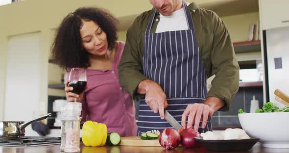 Happy diverse couple preparing food together in kitchen, man chopping vegetables embraced by partner