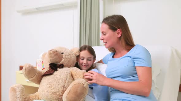 Woman giving a teddy bear to a girl in a bed with balloons