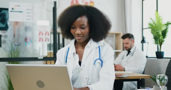 African American Female Doctor in white Coat Sitting at Workplace and Working on Computer