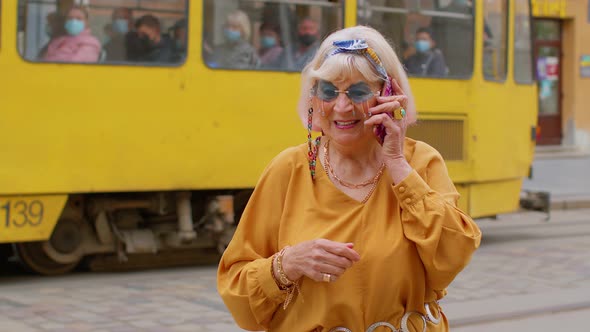 Senior Old Tourist Woman in Stylish Clothes Talking on Mobile Phone While Walking on City Street