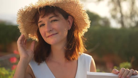 Confident Slim Caucasian Woman Touching Straw Hat Looking at Camera Posing in Sunbeam Outdoors