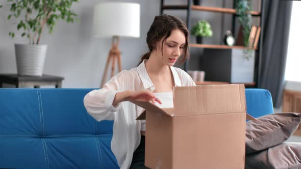 Satisfied Smiling Young Beautiful Woman Looking Into Open Parcel Box at Home on Sofa