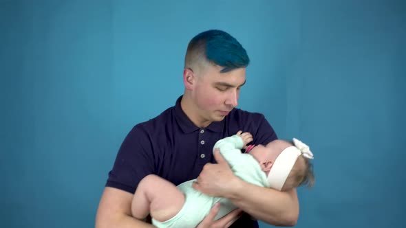 A Young Father with Blue Hair Holds a Baby in His Arms. Alternative Person Cradles a Child on a Blue