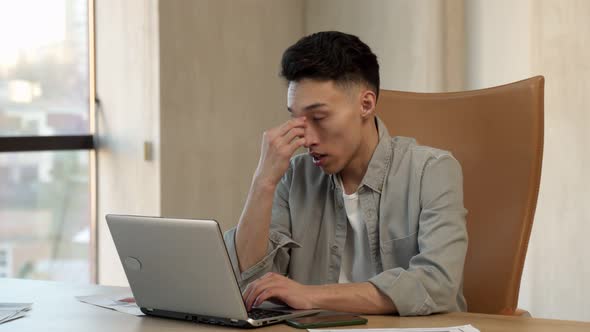 Tired Asian Man Rubs Sleepy Eyes Working on Laptop in Office