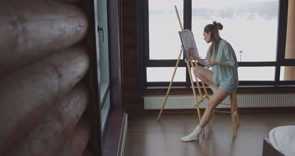 Calm Young Woman Dressed Pajamas is Drawing Picture on Easel Near Window at Home Holding Paintbrush