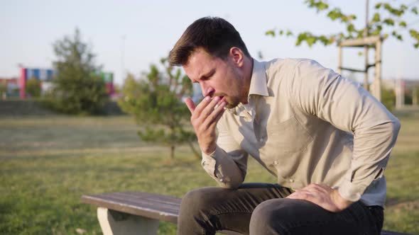 A Young Caucasian Man is Sick Coughs and Has a Headache As He Sits in a Park