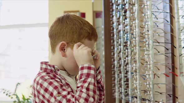 A Redhaired Boy in a Plaid Redandwhite Shirt Tries on Glasses Near a Rack with Lens Frames and