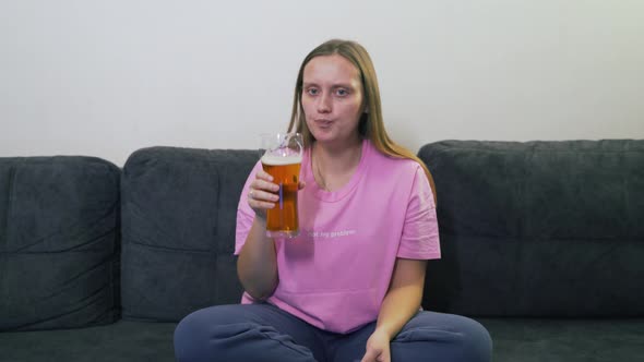 Young Woman is Sitting on Gray Sofa at Home Chewing Food and Drinking Beer From Transparent Glass