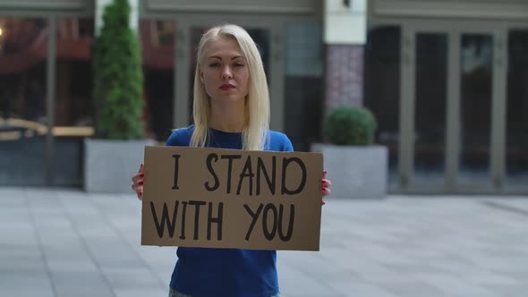 Young Woman Blonde Stands with a Cardboard Poster I STAND WITH YOU in a Public Place Outdoor. Single