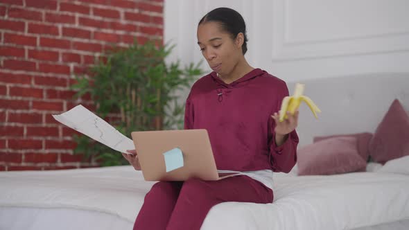 African American Young Woman Eating Banana Analyzing Business Strategy in Bedroom in the Morning