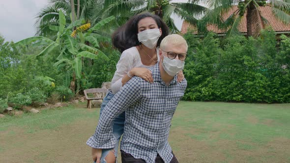 Young Mixed Race Couple Wearing Protective Mask Running Piggyback and Having Fun at the Home Garden