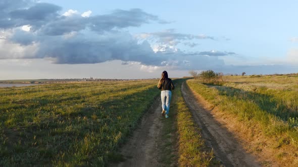 Young Woman Runs on the Field Road Near Water During Beatiful Sunset Aerial Shot