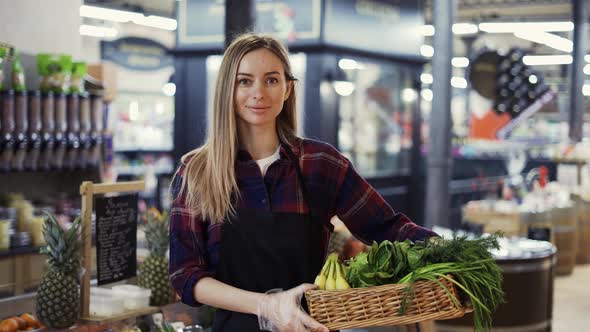 Female Supermarket Employee in Black Apron Holding a Box Full of Fruits and Greens in Supermarket