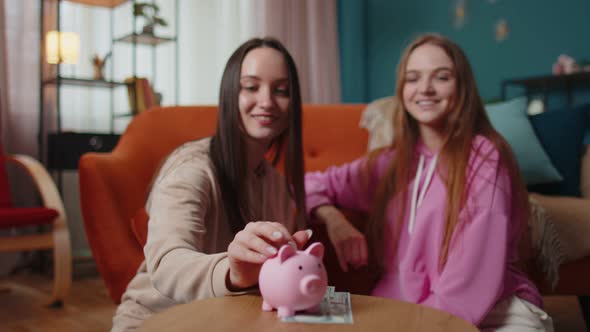 Girls Friends Siblings Sitting on Floor and Take Turns Dropping Dollar Banknote Into Piggy Bank