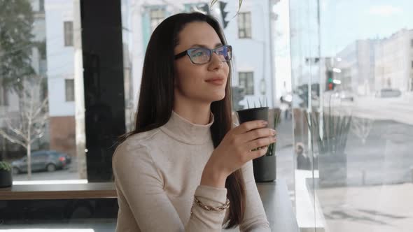 Young Woman with Eyeglasses Drinking Coffee and Watching City Life