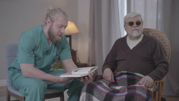 Male Caucasian Volunteer in Nurse Uniform Reading Book To Blind Senior Retiree Indoors. Sympathetic
