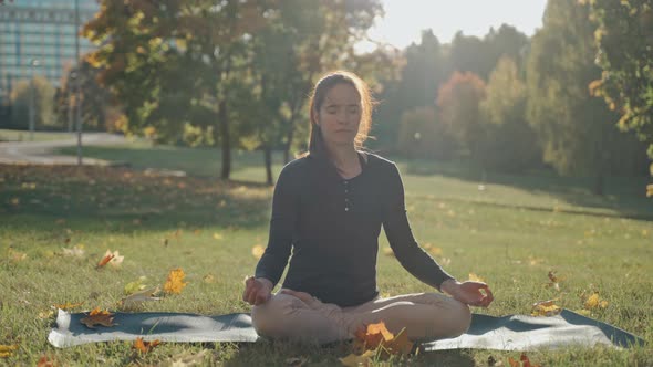 Young Woman Sitting on Mat in Lotus Position in City Park on Sunny Autumn Day