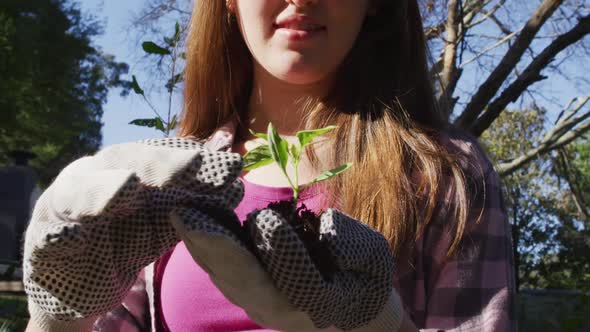 Smiling caucasian teenage girl working in garden wearing gloves and inspecting plants