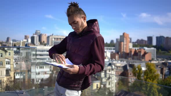 Concentrated African American Man Analyzing Paperwork Standing on Rooftop in Urban City