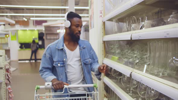 Young African Guy in Headphones with Shopping Cart Choosing Glassware in House Improvement Store