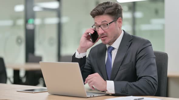 Middle Aged Businessman with Laptop Talking on Smartphone in Office