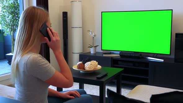 A Blonde Woman Sits on a Couch in a Living Room and Talks on a Smartphone, a TV with a Green Screen