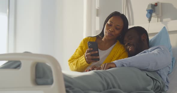 African Couple Resting in Hospital Bed and Using Smartphone Together