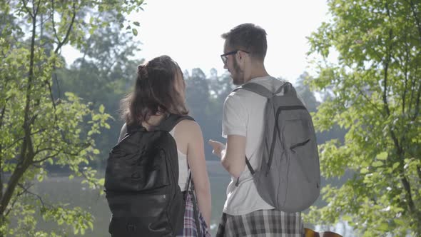 Couple Standing on the Riverbank in the Forest with Backpacks Pointing Away. The Young Man and Woman