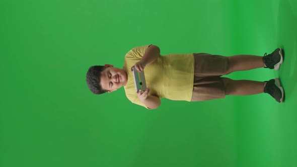 Full Body Of Excited Asian Little Boy Playing Video Game With Mobile Phone In Green Screen Studio