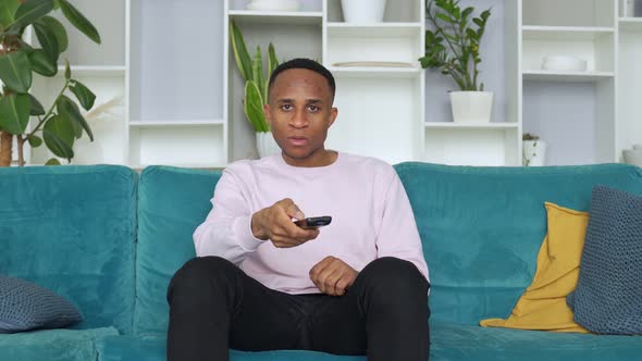 Happy Young African Ethnicity Man Relaxing on Cozy Sofa at Home, Holding Remote Controller, Watching