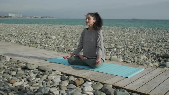 Relaxed Sporty Girl Is Sitting on Wooden Path Near Sea and Meditating